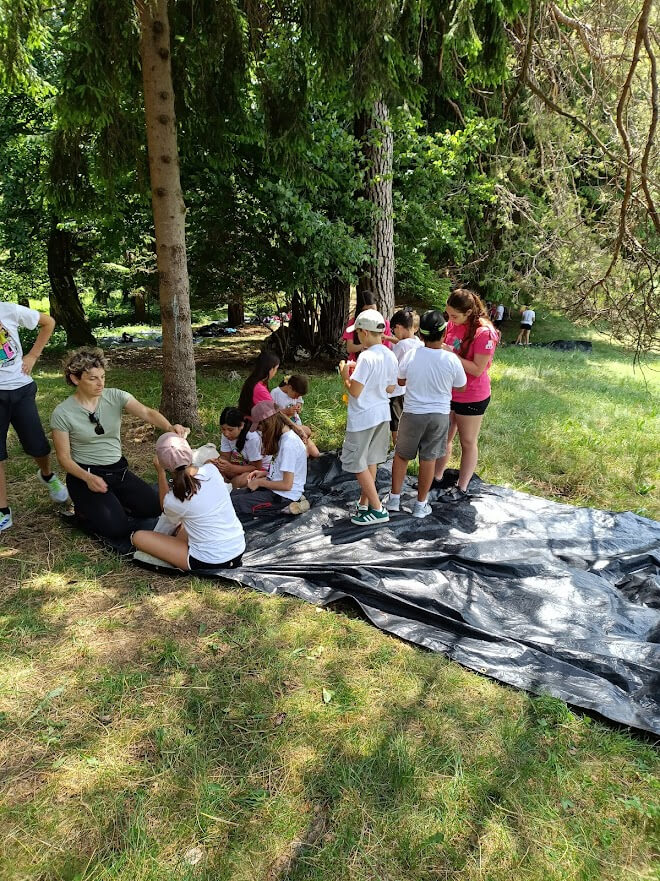 Educatrice con un gruppo di bambini raccolti in cerchio sotto gli alberi durante un momento di spiegazione.