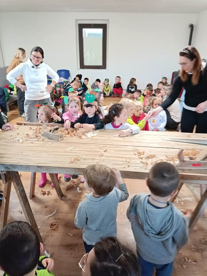 Grande gruppo di bambini raccolto attorno al banco del laboratorio durante una dimostrazione di lavorazione del legno.