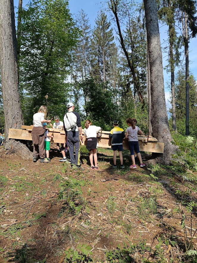 Gruppo di ragazzi attorno a una struttura in legno realizzata all'aperto durante un laboratorio collettivo in foresta.