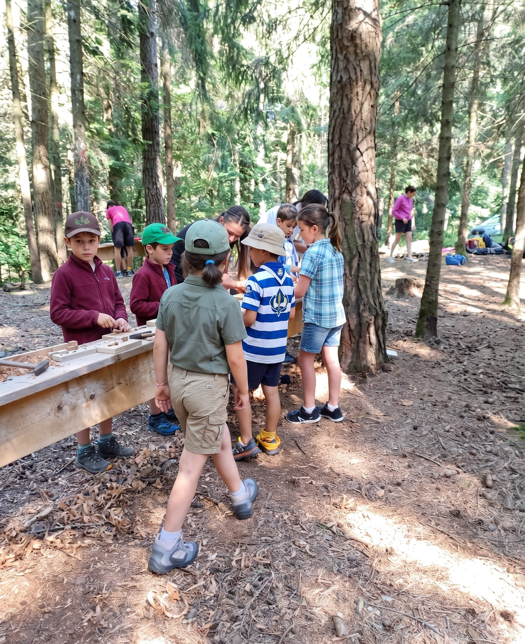 Gruppo di bambini e ragazzi attorno a un tavolo da lavoro nel bosco durante un laboratorio manuale all'aperto.
