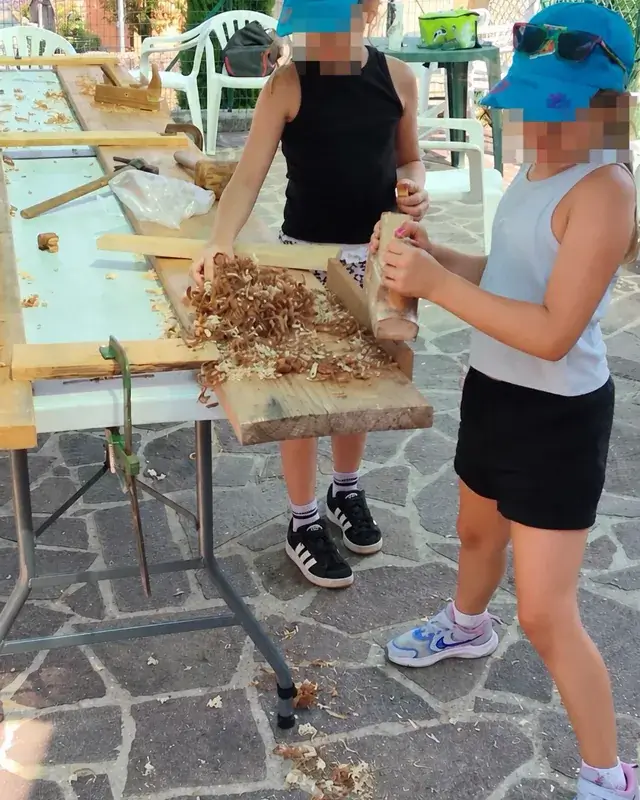 Due bambine piallano il legno lungo il banco all’aperto.