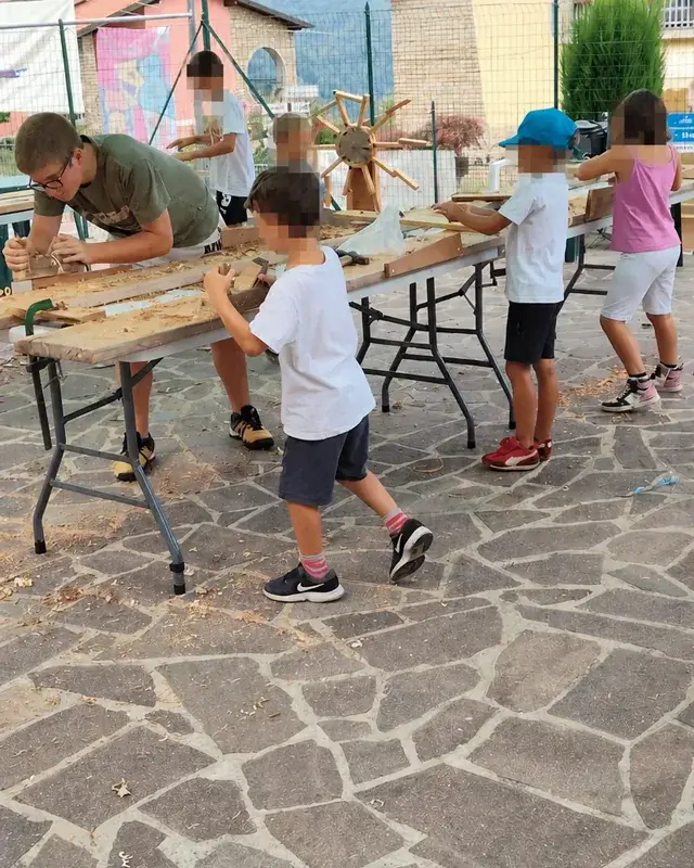 Bambini lavorano a diverse postazioni intorno ai tavoli, tra utensili e trucioli.