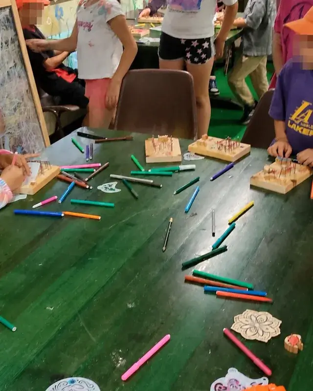Bambini in piedi attorno al tavolo con colori, tavolette e piccoli elementi di legno.
